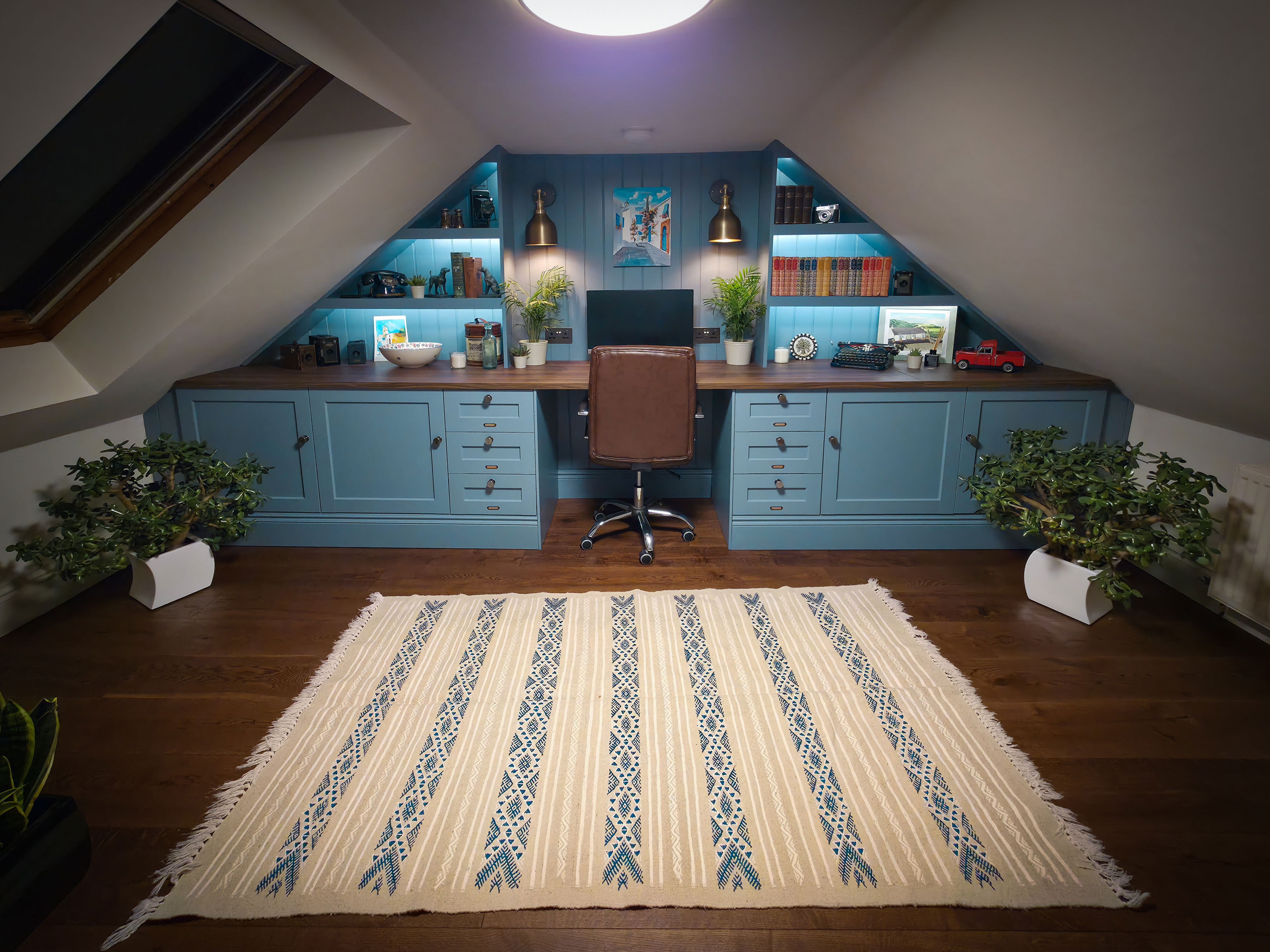 Symmetrical interior view of a modern attic home office featuring deep blue built-in desks and cabinetry, integrated shelf lighting, a brown leather swivel chair, rich dark wood flooring, and a patterned cream area rug under a sloped ceiling with a skylight.