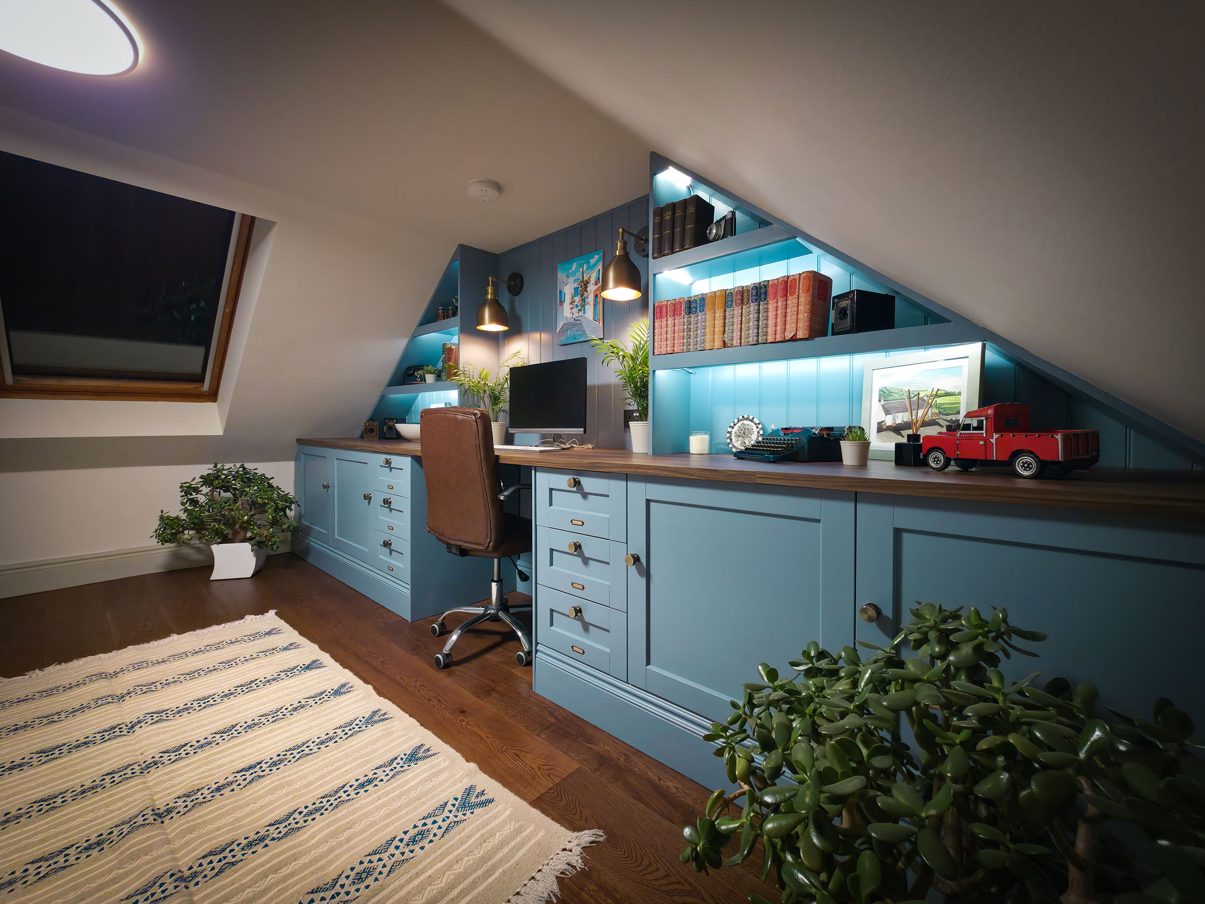 A low-angle, wide interior shot of a bespoke attic office under a sloped ceiling, showcasing built-in blue cabinetry with integrated shelf lighting, a leather office chair, a wooden desk surface with a computer, and a patterned rug on dark wood flooring.