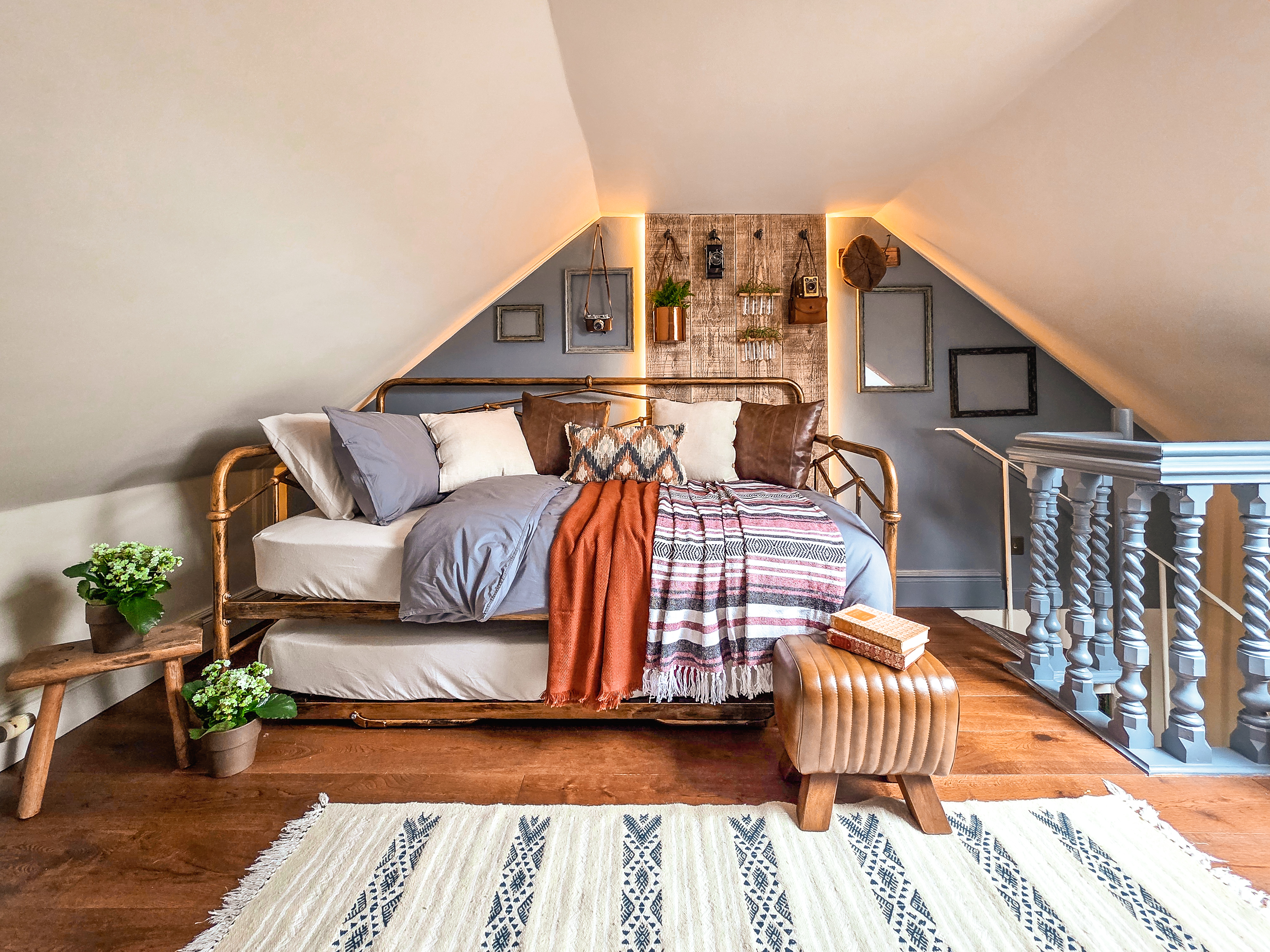 A cozy attic bedroom loft with a rustic metal daybed frame, blue and brown bedding, two dogs sitting on a patterned blanket, a rustic wood accent wall with warm backlight, dark hardwood floors, and a blue painted wooden staircase banister.