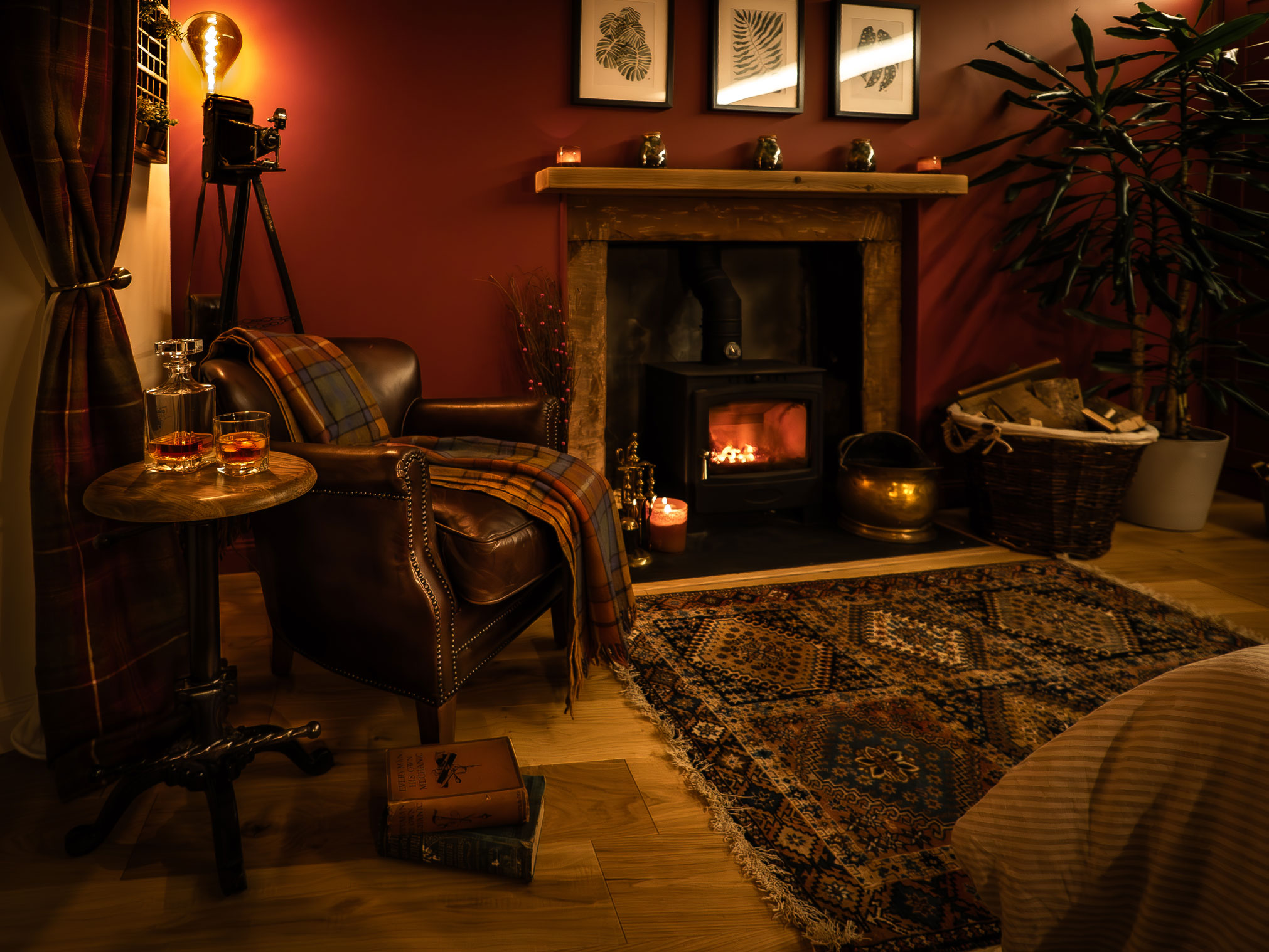 Cozy cottage living room with a roaring black log burner inside a rustic stone fireplace, flanked by a leather armchair with a plaid blanket, a crystal whiskey decanter on a side table, and an oriental rug on wood floors against deep red walls.