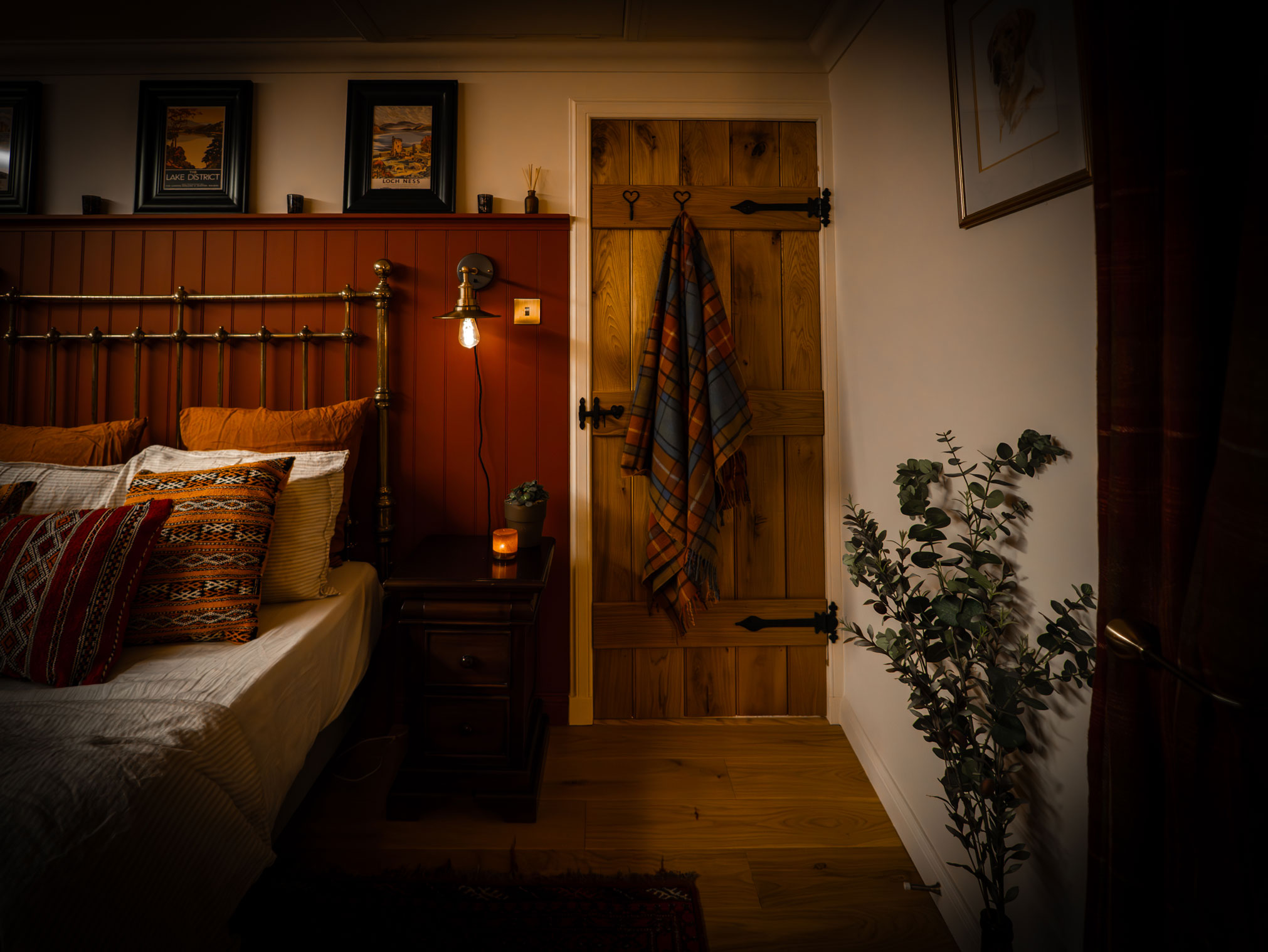 Cozy traditional Scottish cottage bedroom interior featuring a brass bed frame against a terracotta wood-paneled wall, a rustic oak brace-and-ledge door with black wrought iron hinges, a hanging plaid blanket, and warm ambient lighting on hardwood floors.