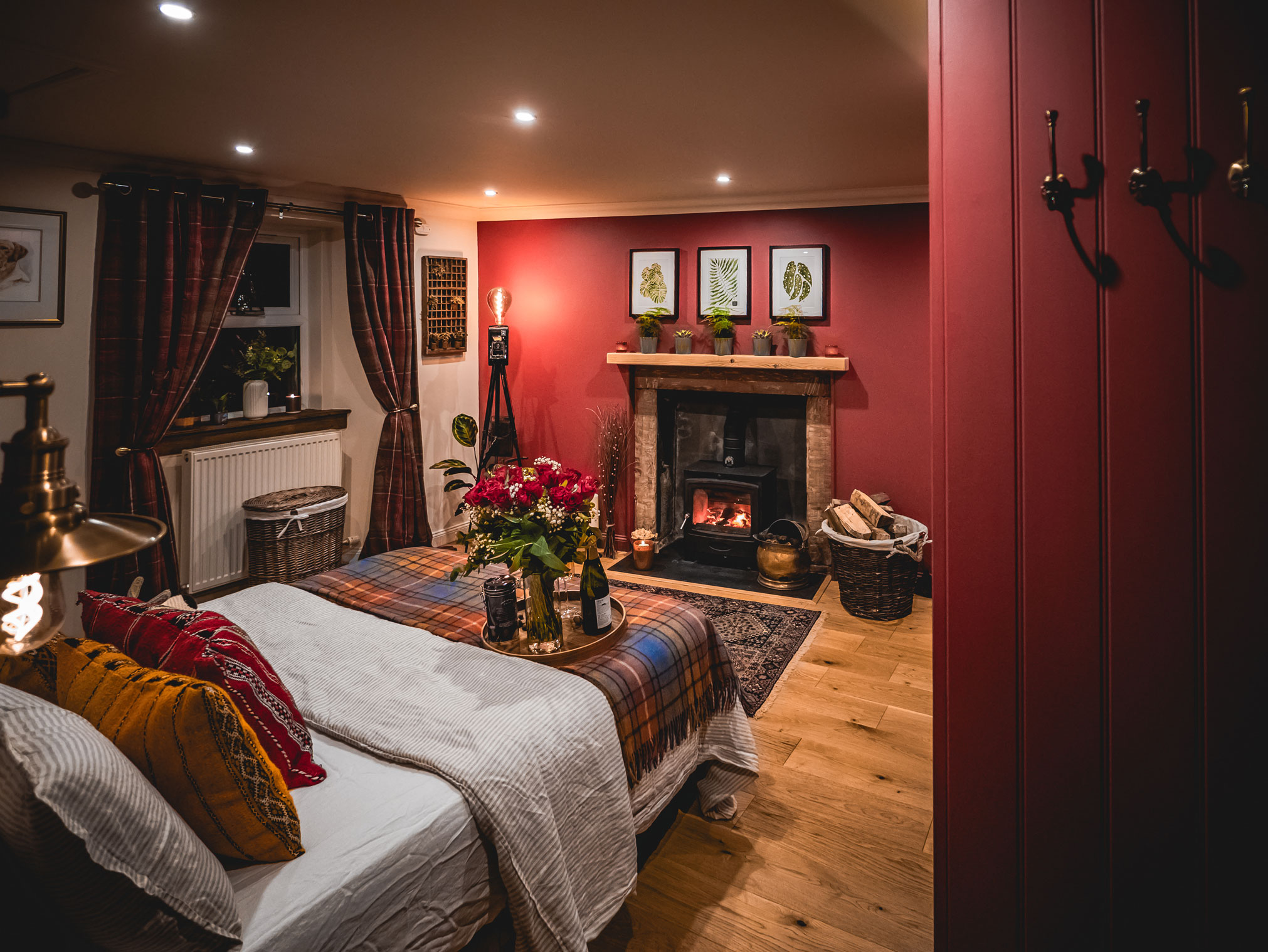 A wide-angle interior view of a cozy cottage bedroom and living space, showing a dressed bed with tartan throws in the foreground and a roaring log burner fireplace against a deep red wall in the background on hardwood floors.