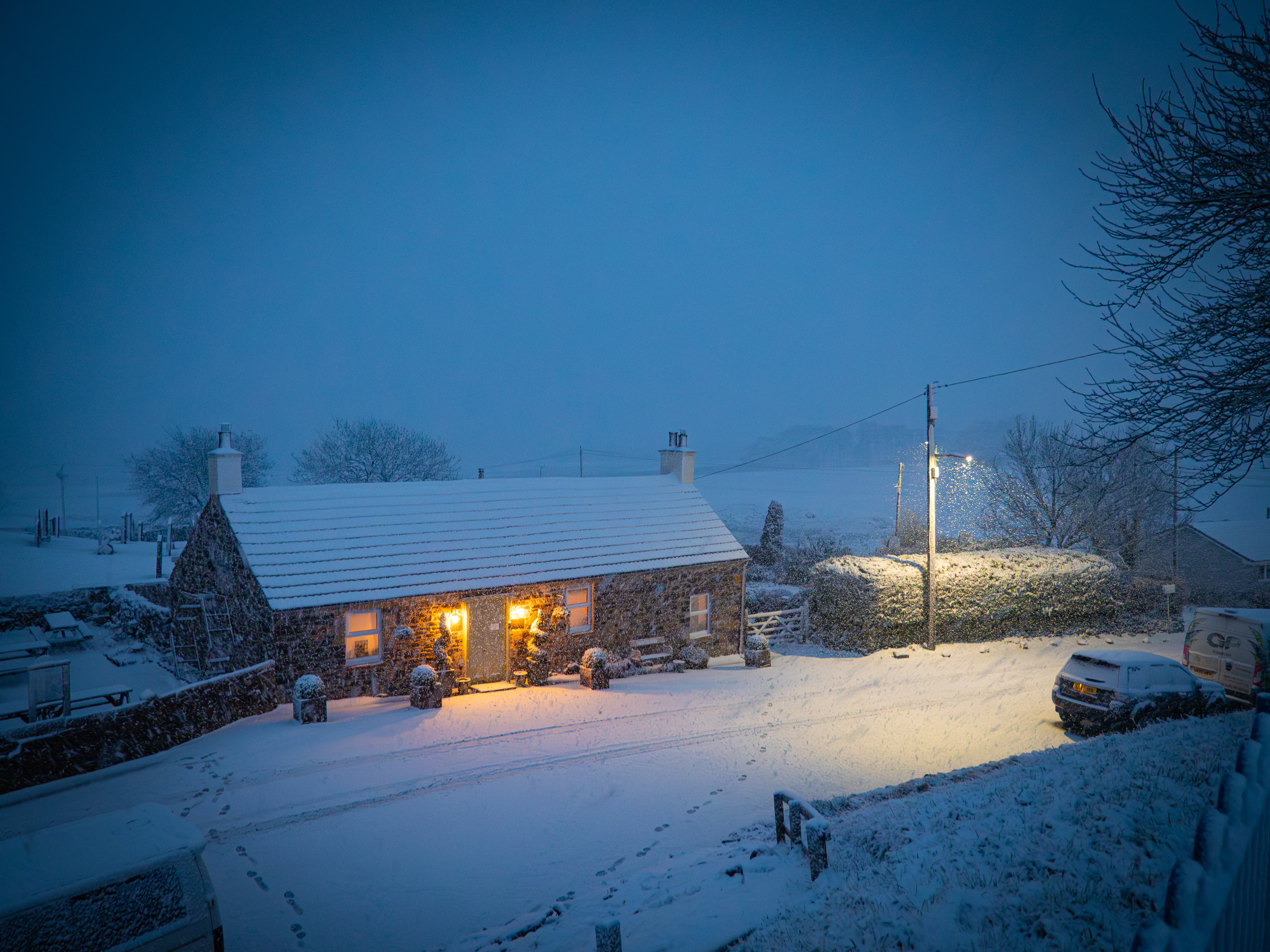 An elevated evening view of a traditional Scottish stone cottage blanketed in thick white snow, with warm glowing windows and coach lights casting a gold light across the snowy driveway under a falling winter twilight sky.