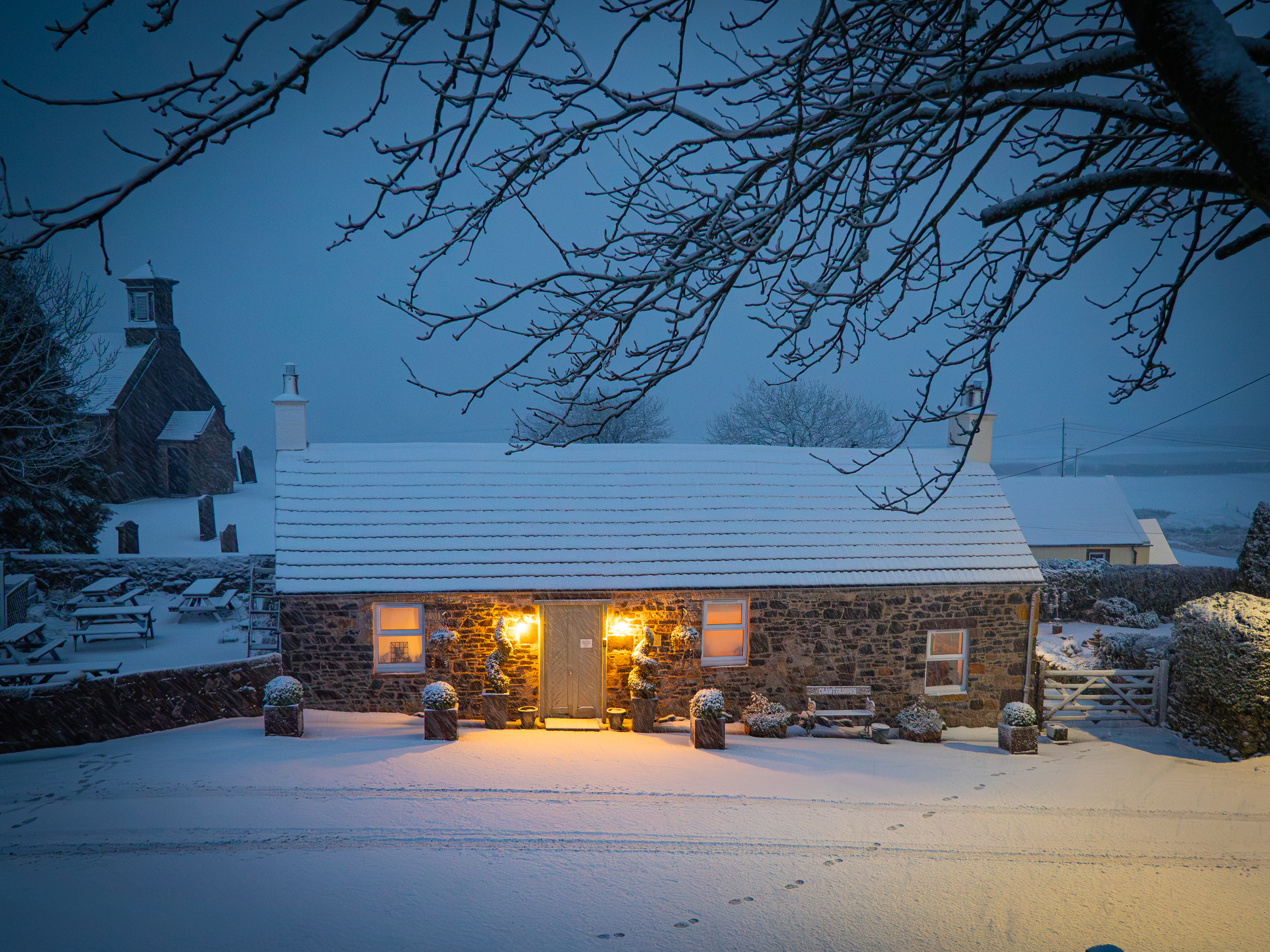A straight-on exterior shot of a stone cottage covered in thick snow at dusk, framed by overhanging snow-dusted tree branches, with glowing warm windows and a historic church tower visible in the background.