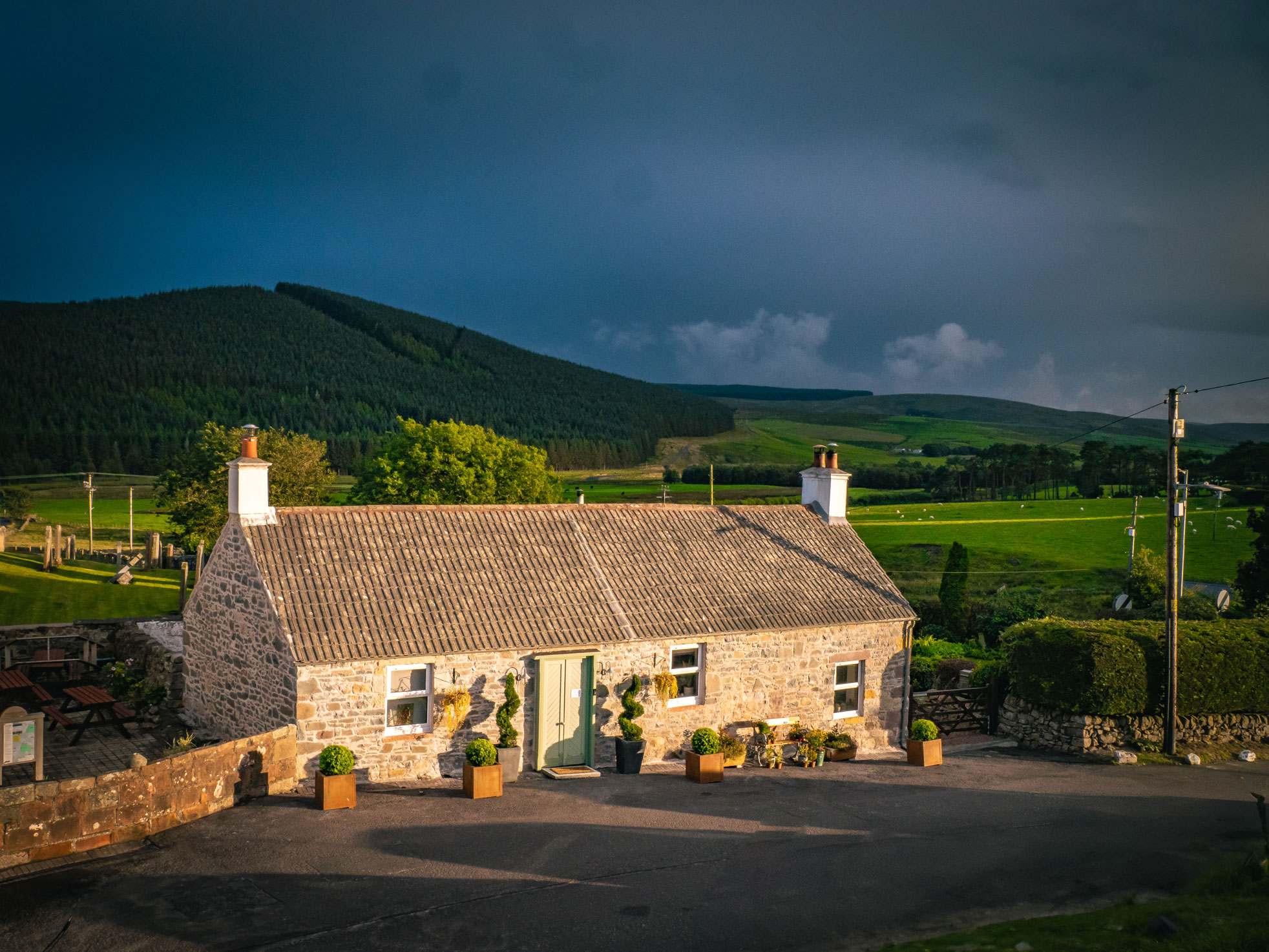 An elevated, wide shot of a traditional single-story stone cottage in Scotland, showing its full length, white chimneys, and sage green door against rolling green hills and a dramatic dark storm sky.