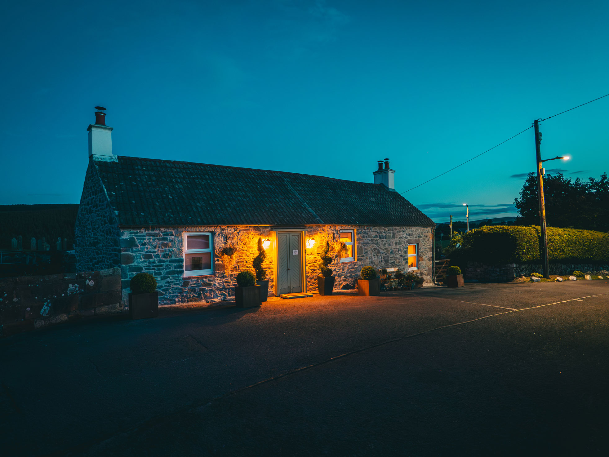 A traditional Scottish stone cottage captured at twilight, featuring glowing warm exterior coach lamps illuminating the stone walls and sage green front door against a deep blue dusk sky.