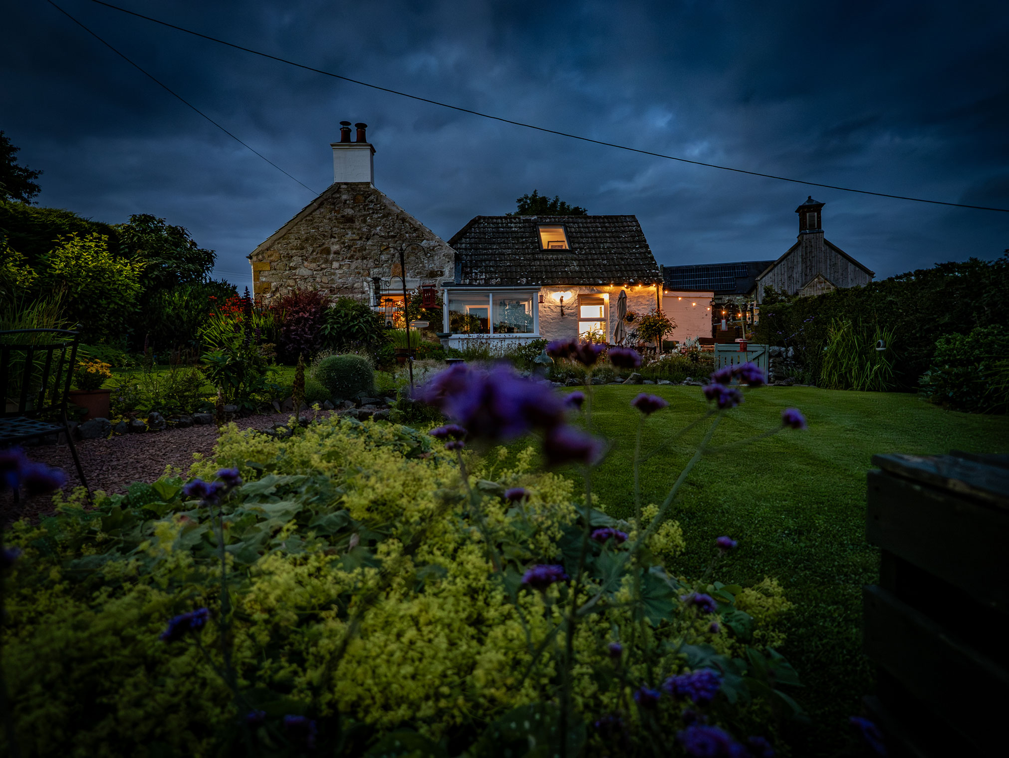 Rear view of Seven Trees Cottage at dusk, showcasing a lush green lawn, landscaped flowerbeds with purple blooms in the foreground, a sunroom extension, and warm interior lights glowing under a moody evening sky.