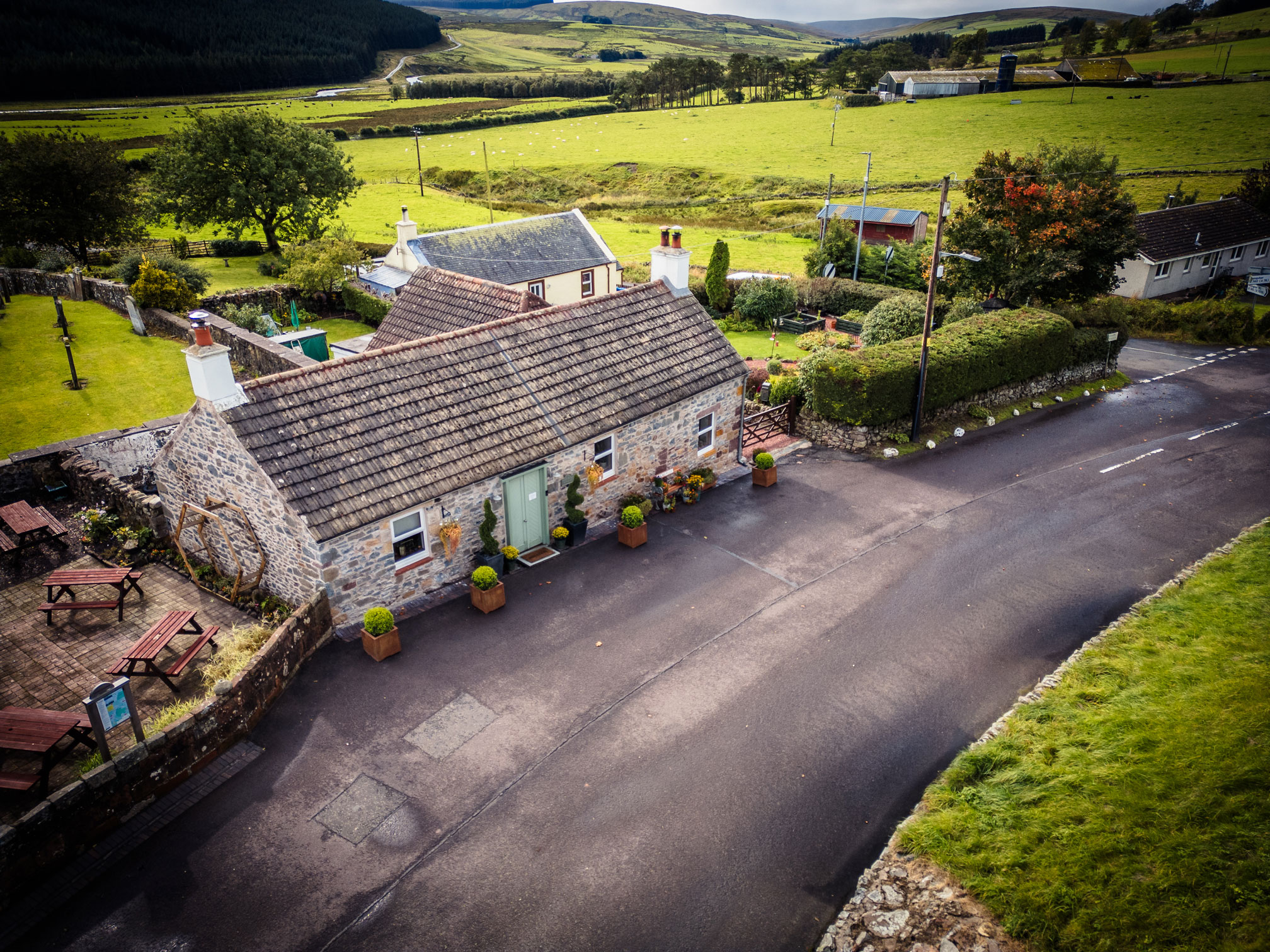 A low aerial view of a traditional stone cottage with a tiled roof and green front door on a quiet country road in Crawfordjohn, Scotland, showing its immediate roadside access and green hills in the distance.