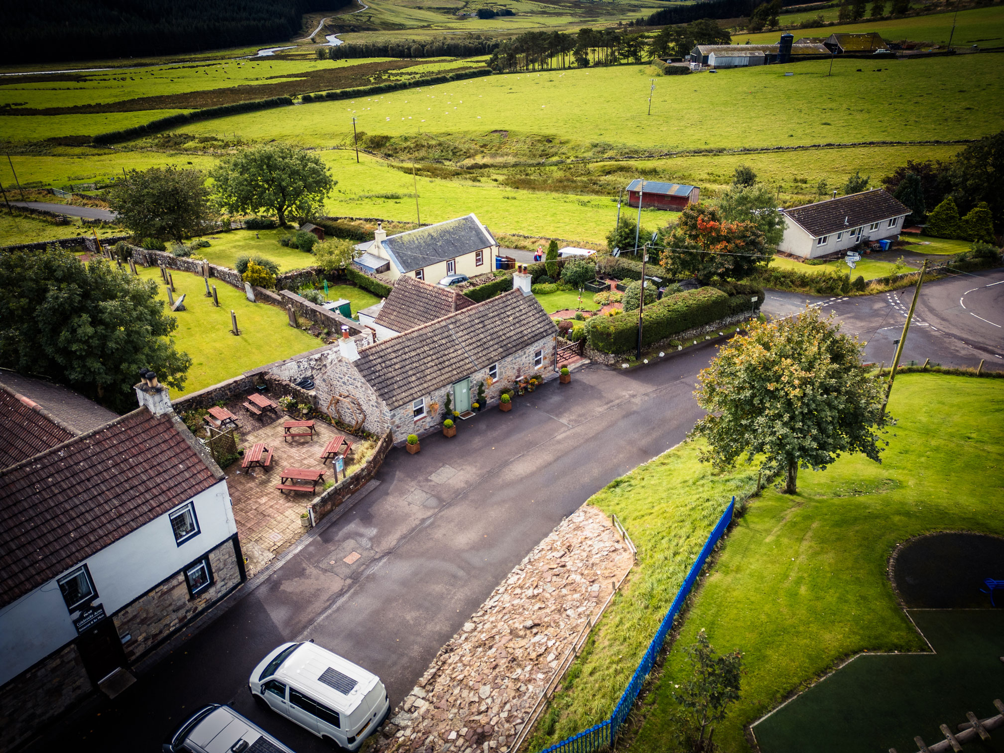 High-angle aerial drone view of Seven Trees Cottage on Main Street in Crawfordjohn, showing the stone cottage, adjacent pub patio with picnic tables, a small historic cemetery, a white van parked nearby, and sweeping green Scottish farmlands stretching into the background.